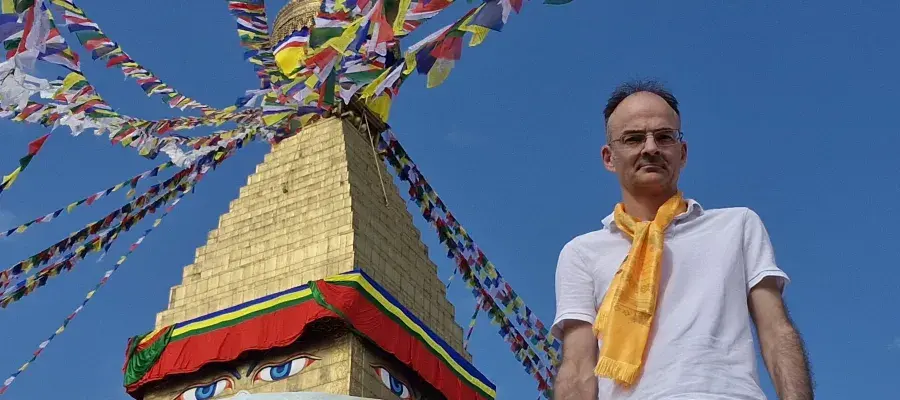 Professor Neil Mabbott, wearing a white T-shirt and yellow scarf, is pictured next to a colourful sculpture against a blue sky.