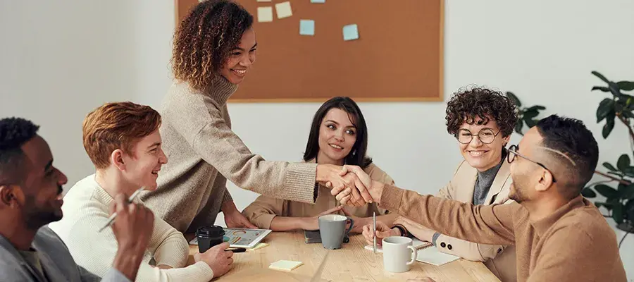 A group of people having a meeting around a table, one person is standing and reaching across the table to shake hands with another person who is seated.