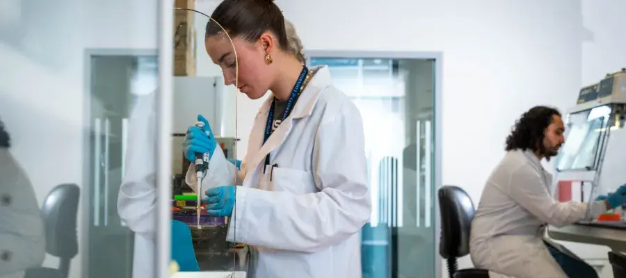 A scientist stands working at a lab bench, while another scientist sits working at the bench opposite. Two scientists in white coats work 