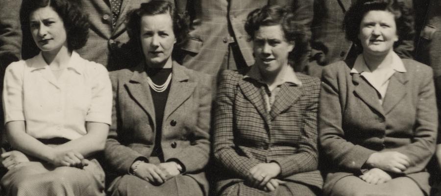 Four women sit in a line, dressed formally. The photo is black and white.