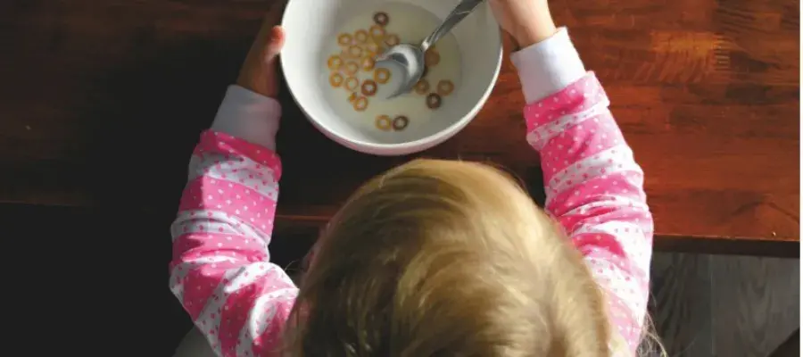 A young child in a pink and white striped top is viewed from above sits at a table eating cereal