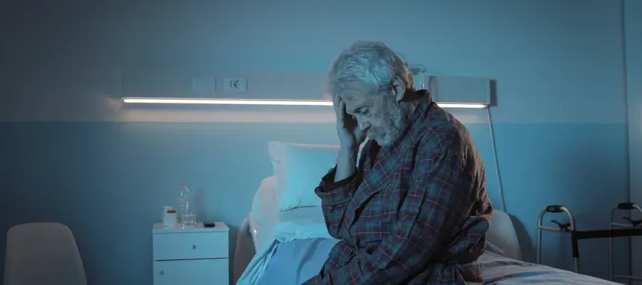 A male patient wearing a checked robe sits on a hospital bed, touching his head