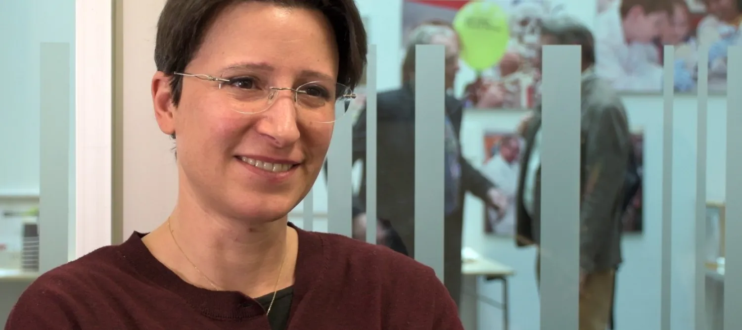 Maria Forlenza, wearing glasses and a dark red cardigan, smiles to someone out of shot