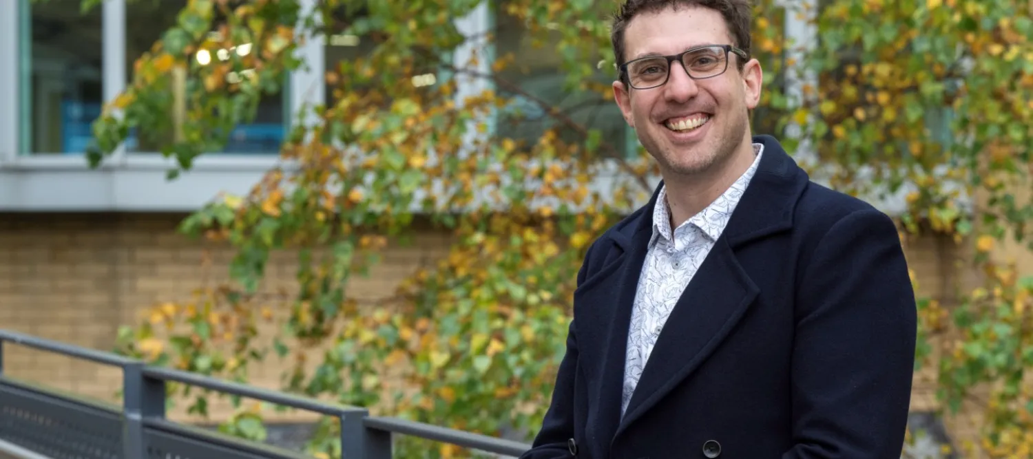 Daniel Tolhurst, wearing a white shirt and navy jacket, is pictured outside a modern office building smiling at the camera