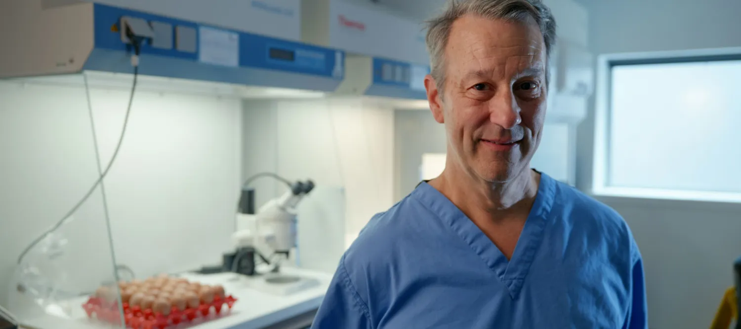 Professor Mike McGrew, dressed in clinical scrubs, stands in front of a fume cupboard in a lab