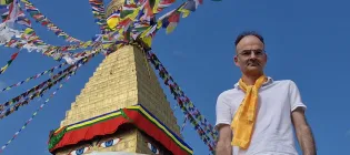Professor Neil Mabbott, wearing a white T-shirt and yellow scarf, is pictured next to a colourful sculpture against a blue sky.