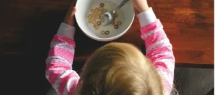 A young child in a pink and white striped top is viewed from above sits at a table eating cereal