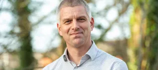 Professor John Hammond, wearing a light-coloured shirt and standing in front of some trees, smiles into the camera.