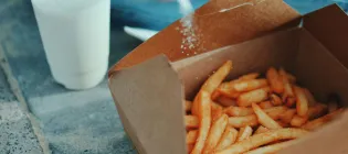 A person shakes salt over a cardboard carton of chips