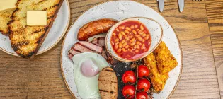A Scottish breakfast on a plate, including a small bowl of baked beans, hash browns, cherry tomatos, a fried egg, sausage, bacon, and black pudding