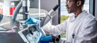 A scientist sits at a stool in front of a microscope in a lab.