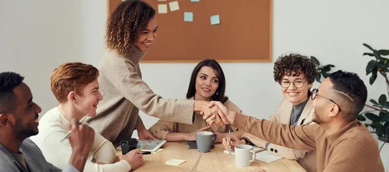 A group of people having a meeting around a table, one person is standing and reaching across the table to shake hands with another person who is seated.