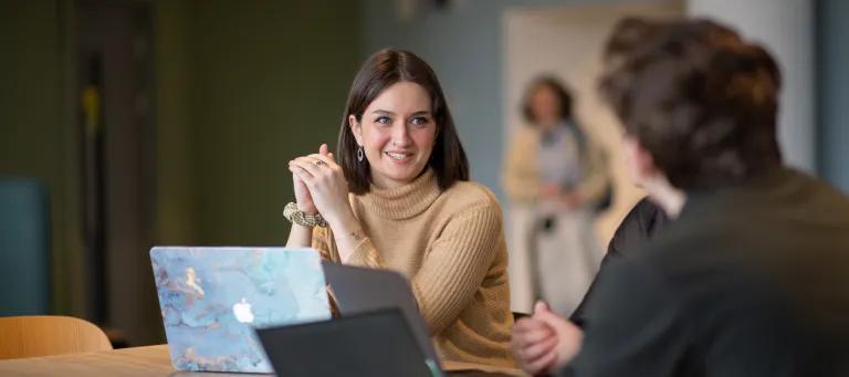 A student with a laptop having a conversation with another student who is sitting in the foreground