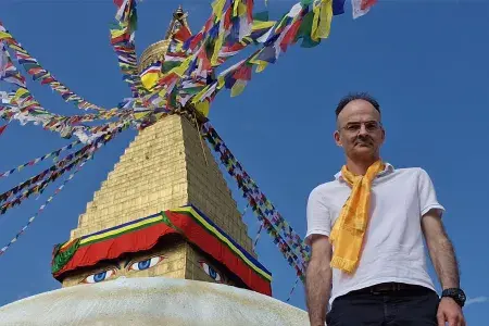 Professor Neil Mabbott, wearing a white T-shirt and yellow scarf, is pictured next to a colourful sculpture against a blue sky.