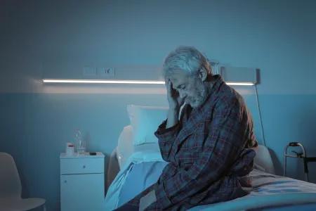 A male patient wearing a checked robe sits on a hospital bed, touching his head