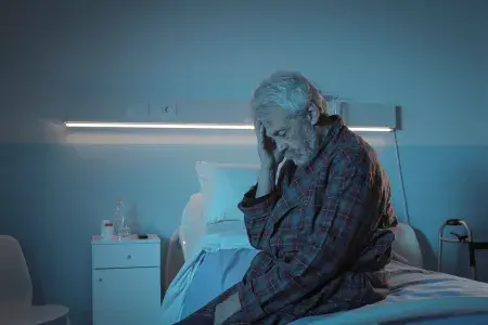 A male patient wearing a checked robe sits on a hospital bed, touching his head