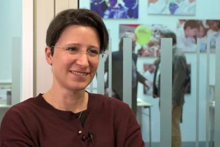 Maria Forlenza, wearing glasses and a dark red cardigan, smiles to someone out of shot