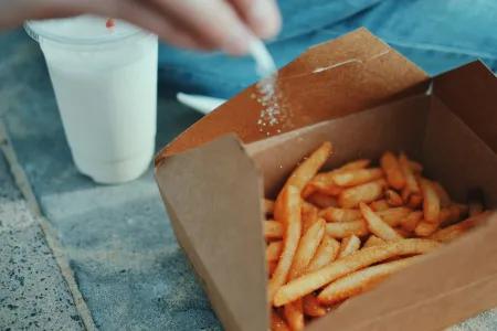 A person shakes salt over a cardboard carton of chips