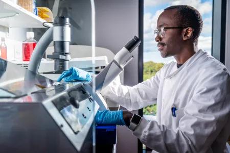 A scientist sits at a stool in front of a microscope in a lab.