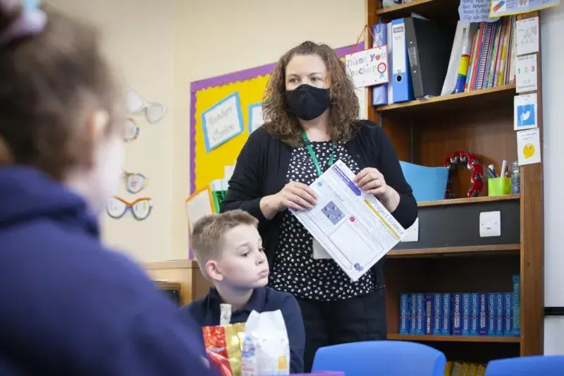 A teacher in a mask holds a worksheet in a classroom