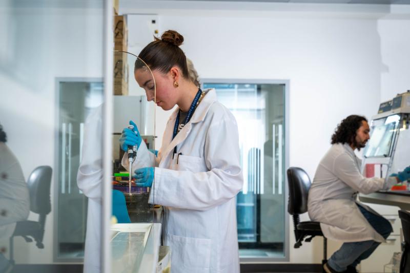 A scientist stands working at a lab bench, while another scientist sits working at the bench opposite. Two scientists in white coats work 