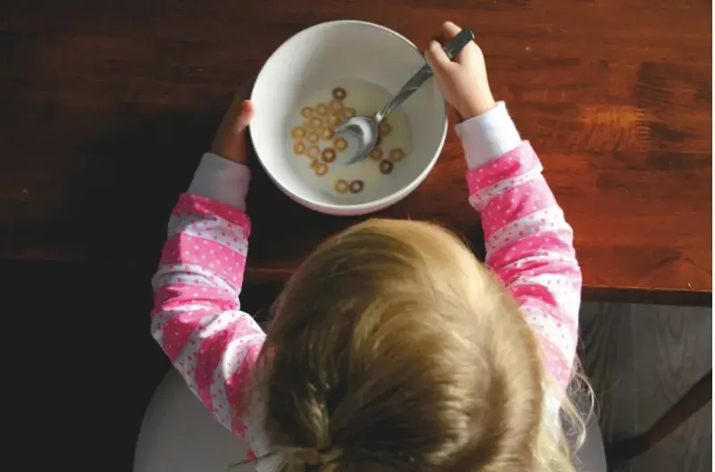 A young child in a pink and white striped top is viewed from above sits at a table eating cereal