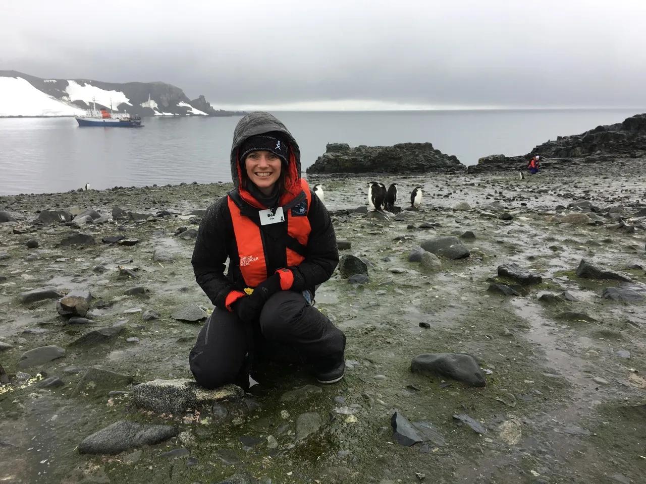 Dr Rebecca Doyle stands smiling in front of a group of penguins in a rocky landscape.
