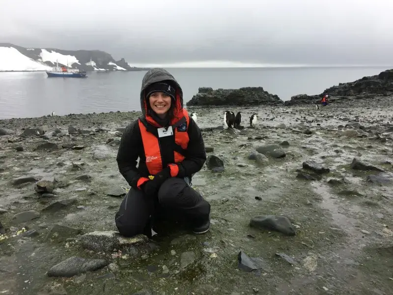 Dr Rebecca Doyle stands smiling in front of a group of penguins in a rocky landscape.