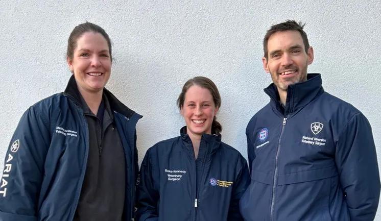 three people stand against a white backfriund, wearing blue outdoor jackets branded Dick Vet Equine Hospital