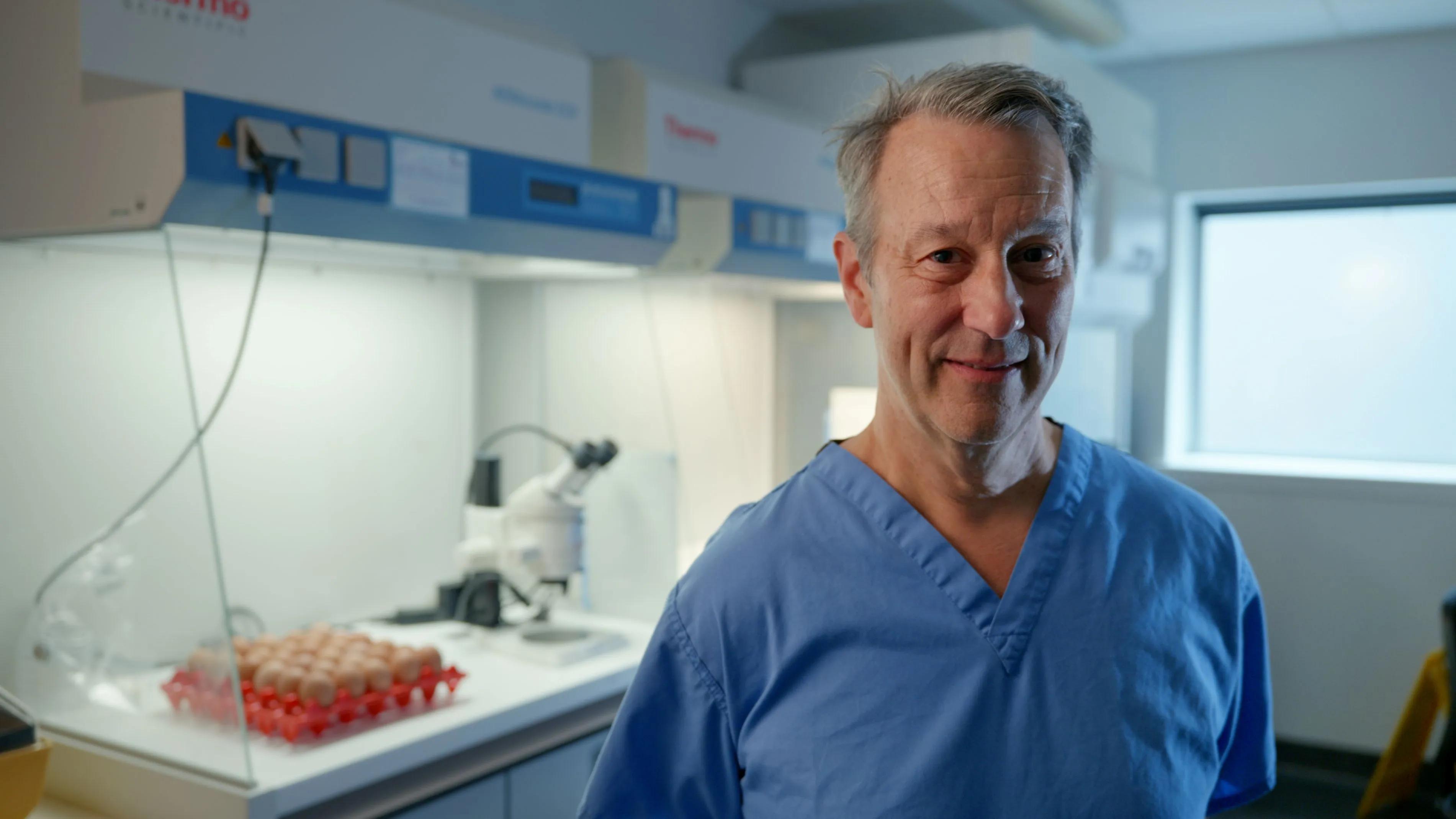 Professor Mike McGrew, dressed in clinical scrubs, stands in front of a fume cupboard in a lab