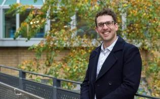 Daniel Tolhurst, wearing a white shirt and navy jacket, is pictured outside a modern office building smiling at the camera