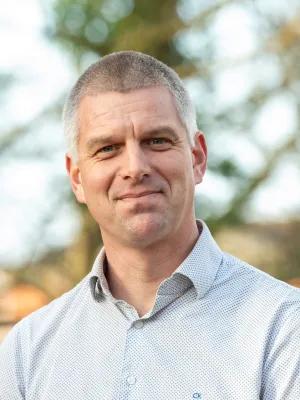 Professor John Hammond, wearing a light-coloured shirt and standing in front of some trees, smiles into the camera.