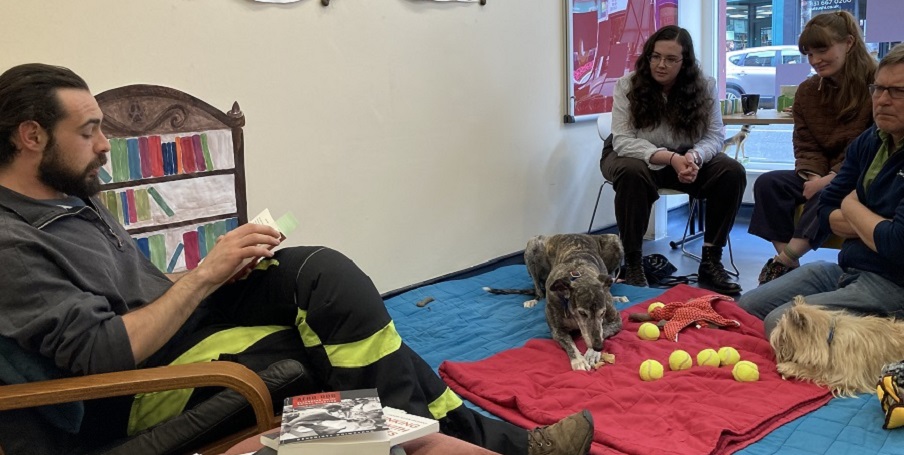 Group of people listen to men reading out loud from a book with dogs at their feet 